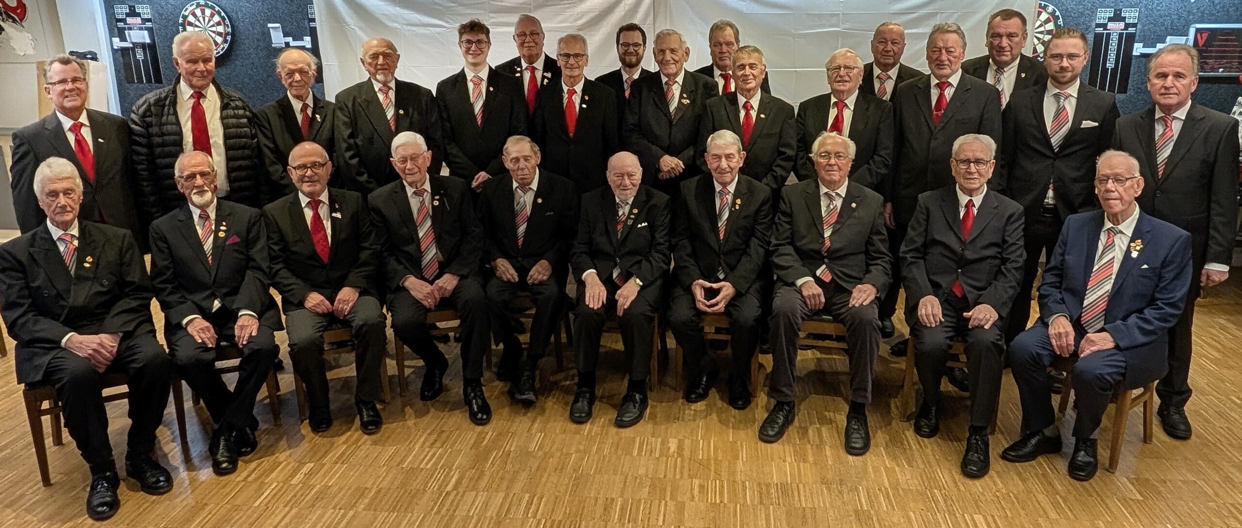 Gruppenfoto der Chorgemeinschaft Q.V. Rheingold-Münster u. MGV Rinkerode in Vereinskleidung: Anzüge mit Vereinskrawatte und Vereinsnadel, aufgenommen im Probenraum unserer Vereinskneipe Mauritius