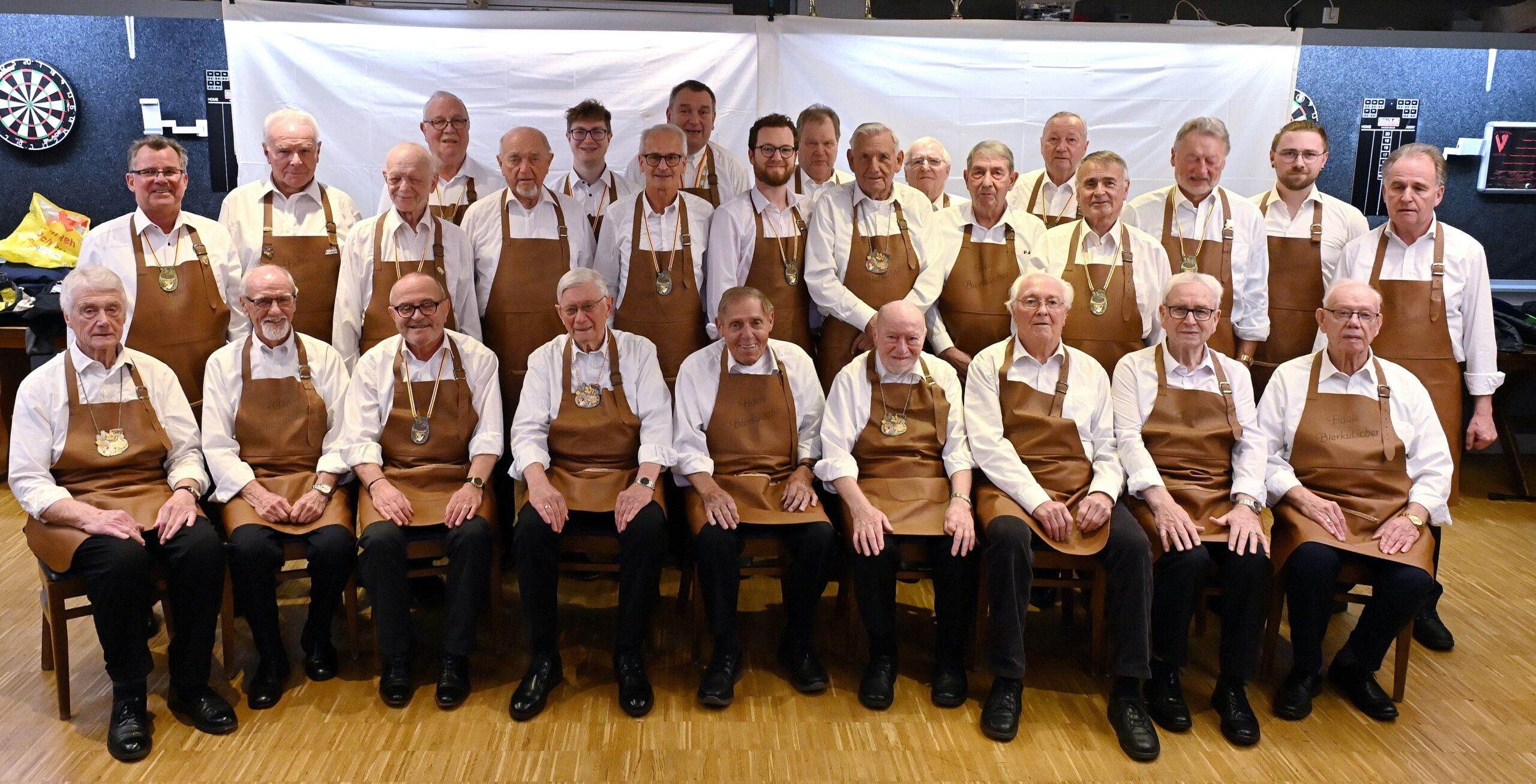 Gruppenfoto der fidelen Bierkutscher in Vereinskleidung: Schlichtes Hemd mit Lederschürze und dem Vereinsorden, aufgenommen im Probenraum unserer Vereinskneipe Mauritius