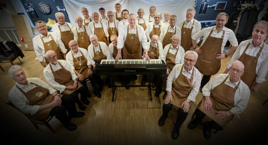 Die fidelen Bierkutscher beim Fotoshooting im Dezember 2024 im Vereinslokal Mauritius. Die Sänger stehen in einer Halbmondform um das Keyboard, an dem Präsident Ulli Schäfers spielt. Sie tragen die neue Vereinskleidung mit weißem Hemd, Lederschürze und dem Orden der fidelen Bierkutscher um den Hals.